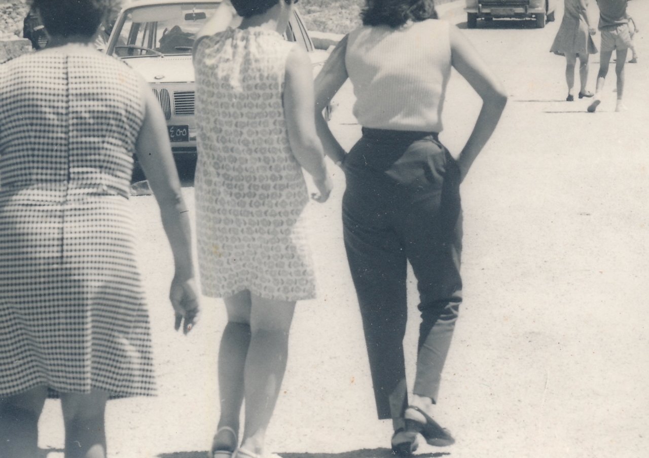 Vintage photo of people walking on a sunny street with classic cars in the background.